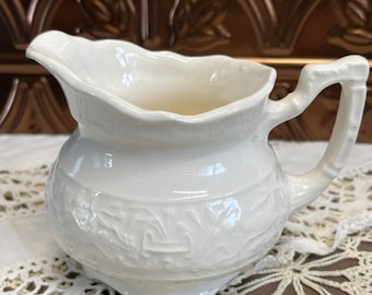 May include: White ceramic creamer with an embossed floral design. The creamer has a rounded body and a handle.