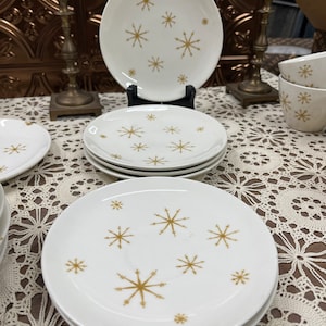May include: White ceramic plates and bowls with a gold starburst pattern. The plates are stacked, and a large plate is displayed on a black stand. Two brass candlesticks are in the background. A set of vintage dinnerware.