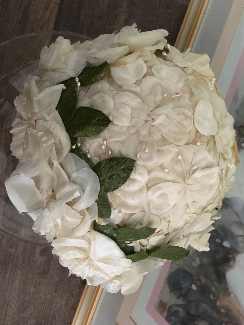 May include: A white fabric hat made of many small flower shapes. The hat is decorated with green leaves and small white beads.