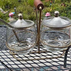 May include: A vintage glass condiment set with two clear glass jars, each with a silver lid and a small spoon. The jars sit in a gold-colored metal holder with a wooden handle. The set is designed for serving condiments.