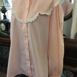 May include: A light pink blouse with a high collar and long sleeves. The blouse features a ruffled collar and cuffs with white lace trim. The front has a button-up closure with silver-colored buttons. The blouse is displayed on a mannequin.