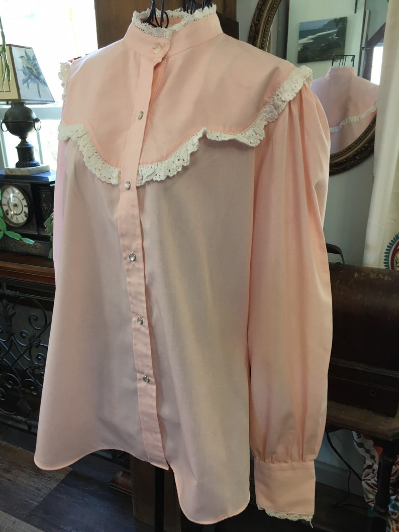 May include: A light pink blouse with a high collar and long sleeves. The blouse features a ruffled collar and cuffs with white lace trim. The front has a button-up closure with silver-colored buttons. The blouse is displayed on a mannequin.