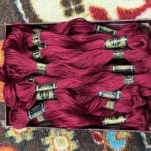May include: A box filled with several skeins of burgundy-colored embroidery floss. Each skein is labeled with a black and gold band, indicating the color and brand. The floss is neatly arranged, suggesting it's ready for crafting projects.