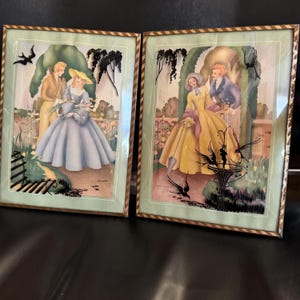May include: Two rectangular framed prints featuring illustrations of couples in period clothing. The left print shows a person in a blue gown and a person in a tan suit. The right print shows a person in a yellow gown and a person in a blue coat.