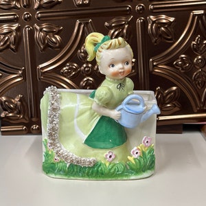 May include: A vintage ceramic planter featuring a young girl in a green dress holding a blue watering can. The planter has a rectangular shape with a floral and grass design, and a decorative silver trim. The girl has blonde hair in a ponytail.