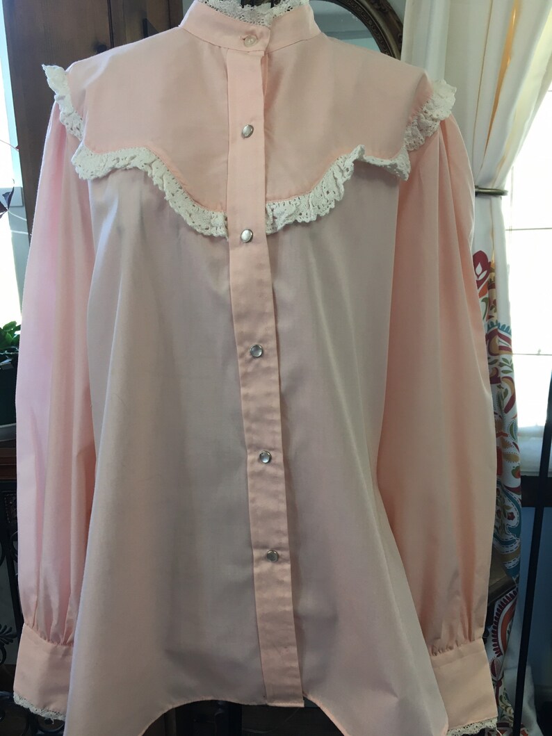 May include: A light pink blouse with a high collar and long sleeves. The blouse features a decorative yoke with white lace trim and silver snap closures down the front. The cuffs also have lace trim.