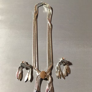 May include: A silver necklace and earrings set with a chain link design and a pendant with a round, silver-toned center. The necklace has a tassel-like design at the bottom with multiple silver-toned leaves.