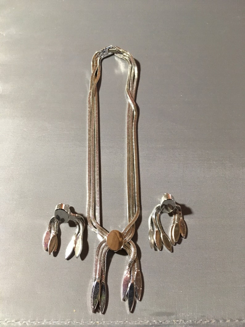 May include: A silver necklace and earrings set with a chain link design and a pendant with a round, silver-toned center. The necklace has a tassel-like design at the bottom with multiple silver-toned leaves.