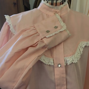 May include: A light pink blouse with a high collar and long sleeves. The blouse features white lace trim along the collar, cuffs, and yoke. Silver-colored snap closures adorn the front and cuffs. The sleeves are gathered at the cuffs.