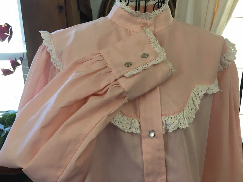 May include: A light pink blouse with a high collar and long sleeves. The blouse features white lace trim along the collar, cuffs, and yoke. Silver-colored snap closures adorn the front and cuffs. The sleeves are gathered at the cuffs.