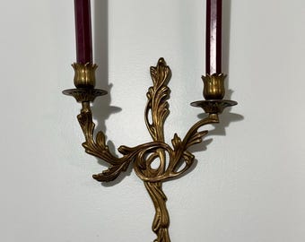 May include: A brass wall sconce with two candle holders, each holding a burgundy-colored candle. The sconce has an ornate, floral design. The candles are approximately 8 inches tall.