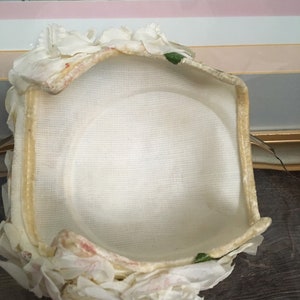 May include: A white fabric hat with a ruffled trim and green leaves. The hat is lined with a white fabric and has a cream colored brim.