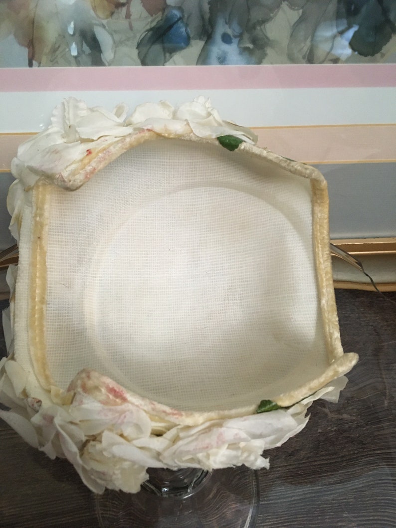 May include: A white fabric hat with a ruffled trim and green leaves. The hat is lined with a white fabric and has a cream colored brim.