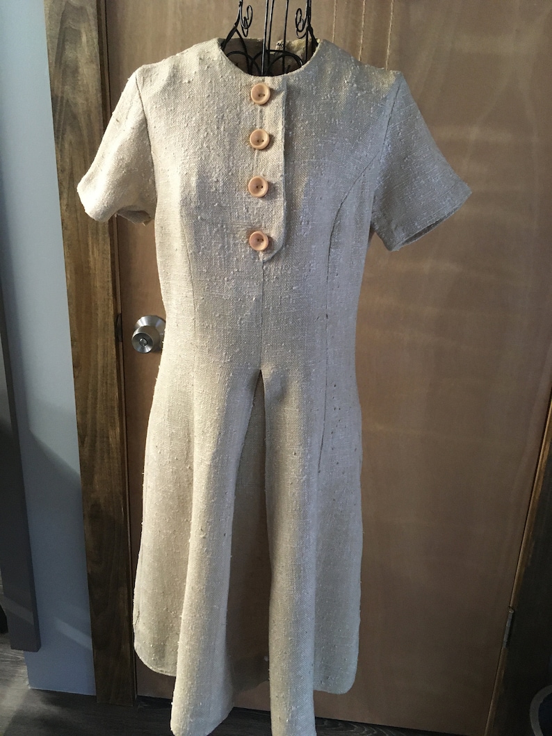 Puede incluir: Un vestido de lino beige con botones en la parte delantera y mangas cortas. El vestido tiene un corpi&ntilde;o ajustado y una falda ligeramente acampanada.