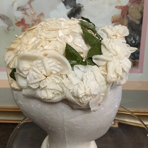 May include: A white hat made of fabric flowers with green leaves. The hat is on a white mannequin head.