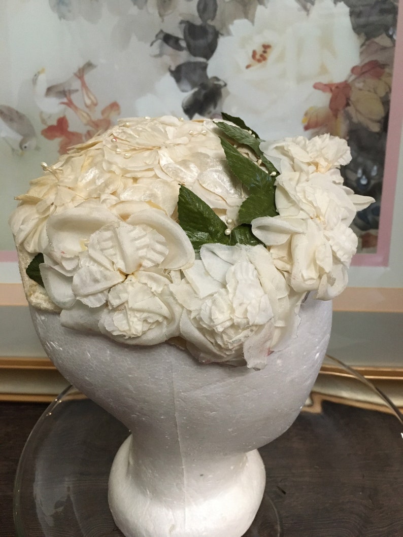 May include: A white hat made of fabric flowers with green leaves. The hat is on a white mannequin head.