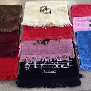 May include: A collection of rectangular terry cloth towels with fringed edges, featuring various embroidered designs including dancing figures, musical notes, geometric shapes, and the word Dancing in white thread across a dark blue towel.
