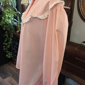 May include: A light pink blouse with a high collar and long sleeves. The collar and cuffs are trimmed with white lace. The blouse has a loose, flowing fit and is made of a soft fabric.