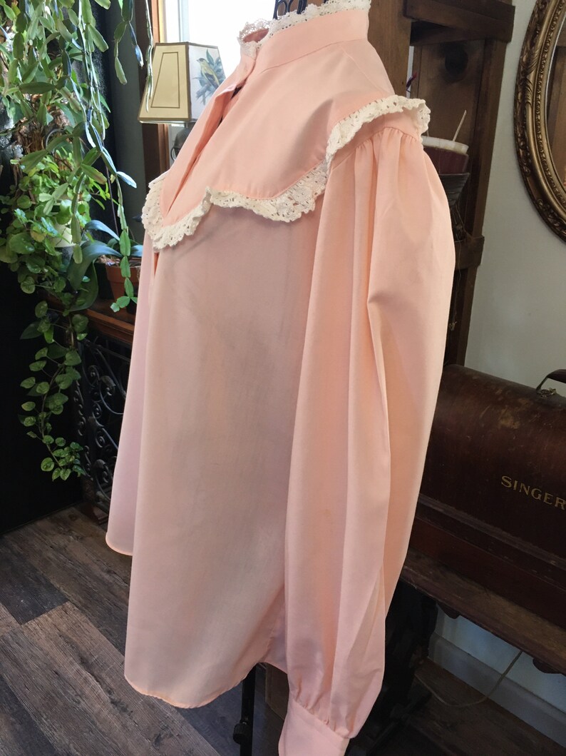May include: A light pink blouse with a high collar and long sleeves. The collar and cuffs are trimmed with white lace. The blouse has a loose, flowing fit and is made of a soft fabric.
