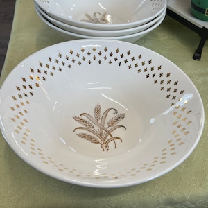 May include: A set of four white ceramic bowls. Each bowl features a gold wheat design in the center and a gold fleur-de-lis pattern around the rim. The bowls are on a light green tablecloth.