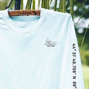 May include: Light blue long-sleeve shirt with a boat graphic and the word "CAPTAIN". The sleeve has the coordinates "44&deg; 31' 48.756" N 69&deg;" printed in black. The shirt is hanging on a wooden hanger.