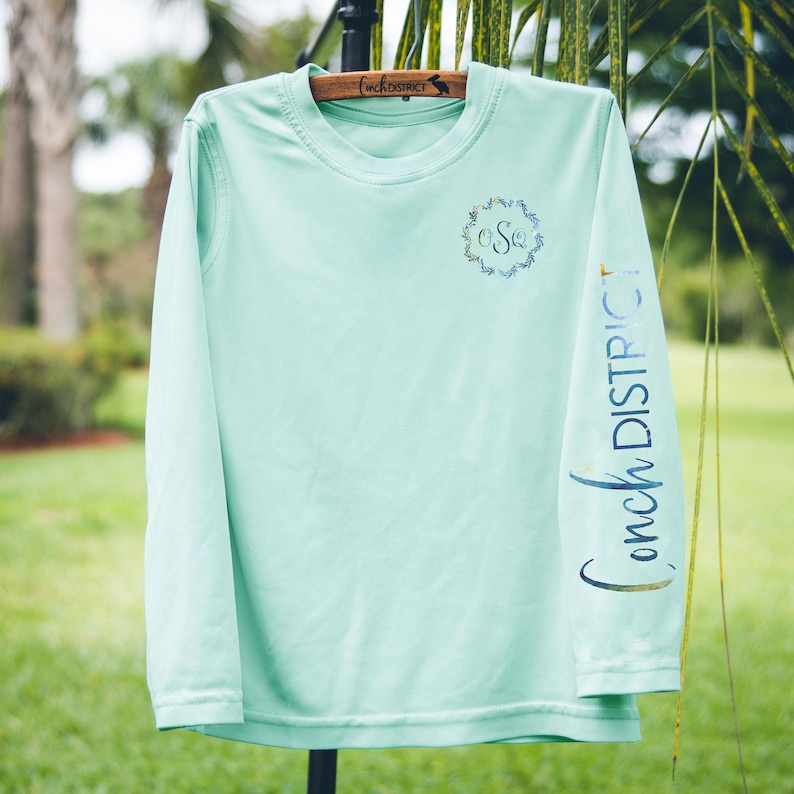 May include: A light green long-sleeved shirt with a floral wreath monogram on the front and the text "Conch District" on the sleeve.