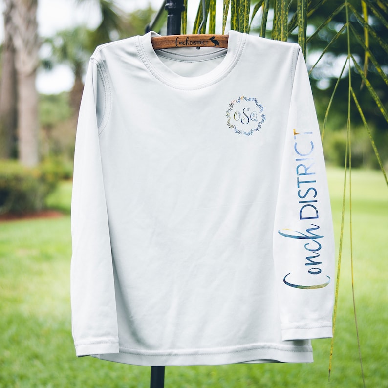 May include: White long-sleeved shirt with a floral monogram and "Conch District" printed on the sleeve.