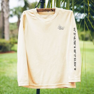May include: A long-sleeved, light tan shirt with a crew neck. The shirt features a small graphic of a ship with the word "CAPTAIN" below it. Coordinates are printed vertically on the right sleeve: 44&deg; 31' 48.756" N 69&deg; 17' 53.304" W.