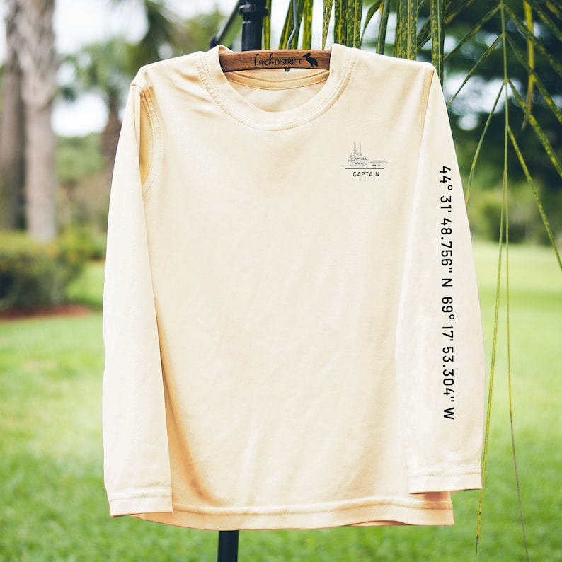 May include: A long-sleeved, light tan shirt with a crew neck. The shirt features a small graphic of a ship with the word "CAPTAIN" below it. Coordinates are printed vertically on the right sleeve: 44&deg; 31' 48.756" N 69&deg; 17' 53.304" W.