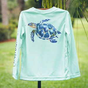 May include: A light blue long-sleeved shirt with a sea turtle graphic on the back. The turtle is painted in shades of blue, green, and yellow.