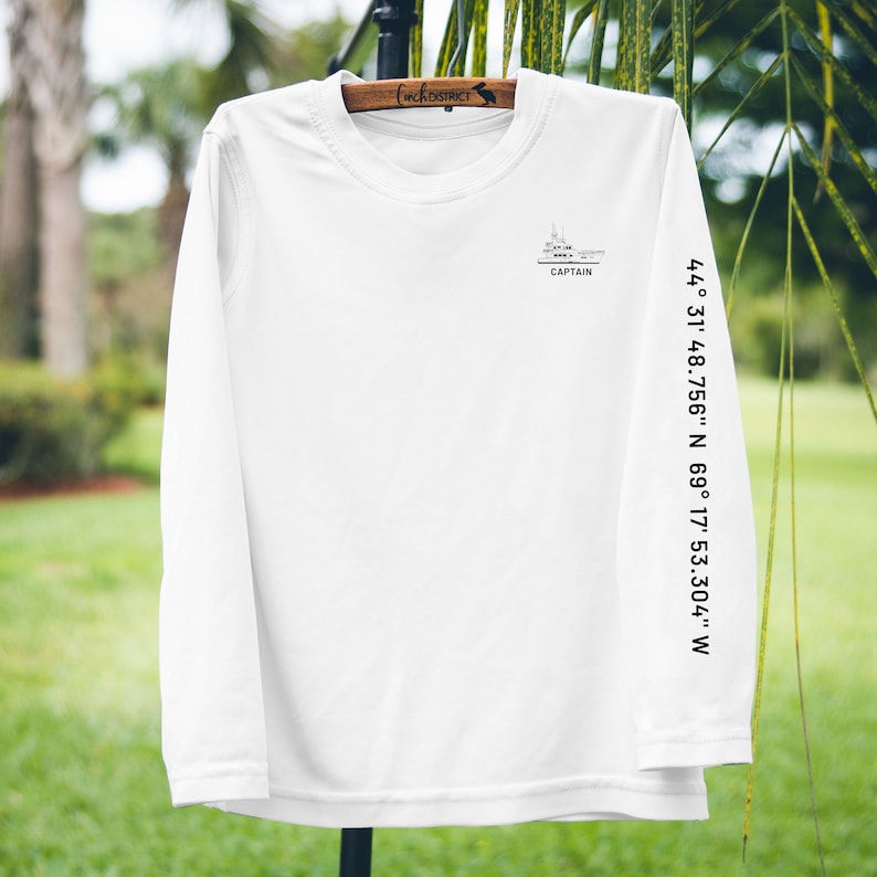 May include: White long-sleeve shirt with a boat graphic and the word "CAPTAIN" printed below it. Coordinates are printed on the sleeve: 44&deg; 31' 48.756" N 69&deg; 17' 53.304" W. The shirt is hanging on a wooden hanger.