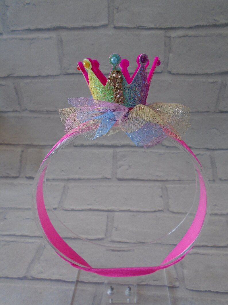 Puede incluir: Una diadema rosa con una corona de purpurina arco&iacute;ris y un lazo de tul. La corona tiene purpurina rosa, amarilla, azul y verde.