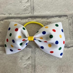 May include: A white hair bow with a yellow elastic band. The bow is covered in white polka dots with red, blue, green, and yellow colors.