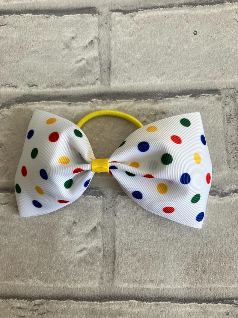 May include: A white hair bow with a yellow elastic band. The bow is covered in white polka dots with red, blue, green, and yellow colors.