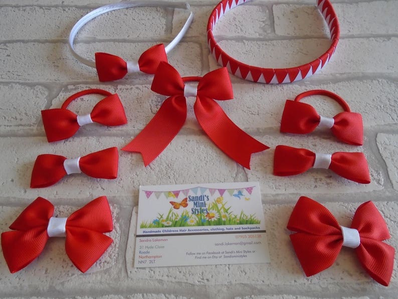 May include: A collection of red hair accessories, including headbands, bows, and ponytail holders, with white accents. The accessories are arranged on a light-colored brick background. A business card is also present.