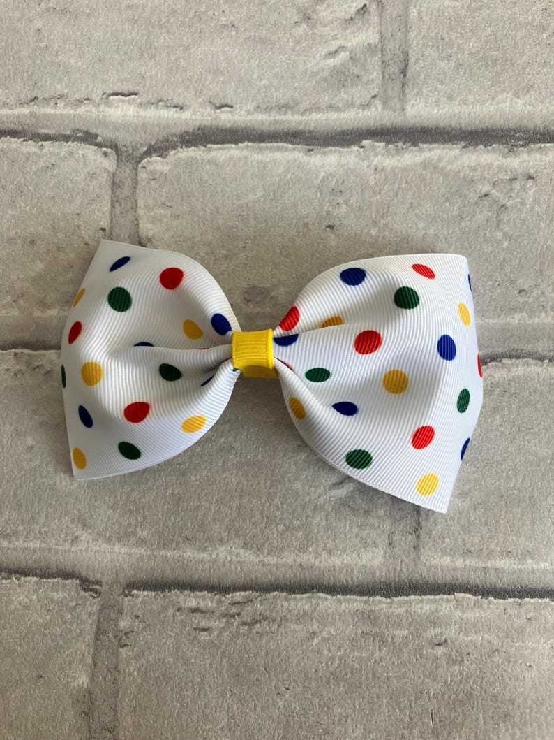 May include: A white bow with red, yellow, blue, and green polka dots. The bow is tied with a yellow ribbon.