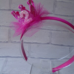 Puede incluir: Diadema rosa con un lazo de tul rosa y una corona rosa brillante con el n&uacute;mero "2" en ella.