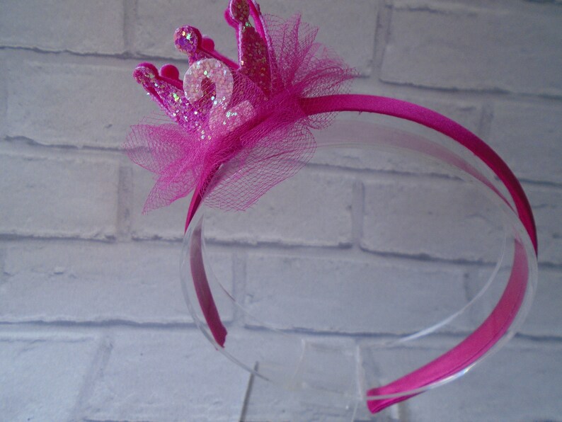 Puede incluir: Diadema rosa con un lazo de tul rosa y una corona rosa brillante con el n&uacute;mero "2" en ella.