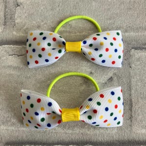May include: Two white hair ties with yellow centers and a colorful polka dot pattern. The hair ties are made of ribbon and have a bright green elastic band.