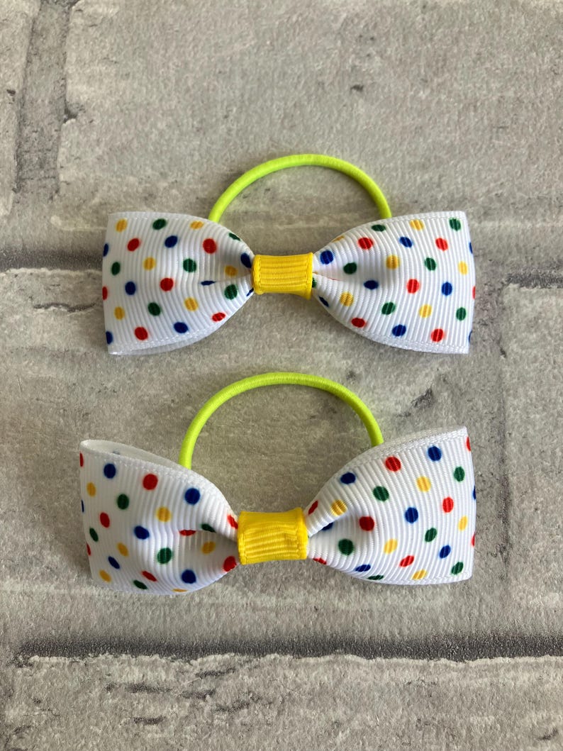 May include: Two white hair ties with yellow centers and a colorful polka dot pattern. The hair ties are made of ribbon and have a bright green elastic band.