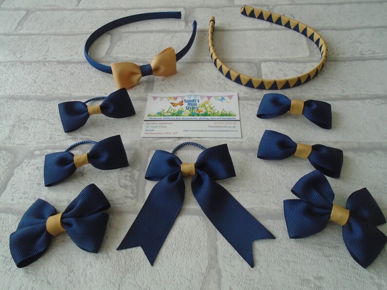 May include: A collection of navy blue and gold hair accessories, including headbands and bow-tie hair ties. One headband is navy blue with a gold bow, and the other is gold with navy blue triangles. The hair ties have a navy blue bow with a gold center.