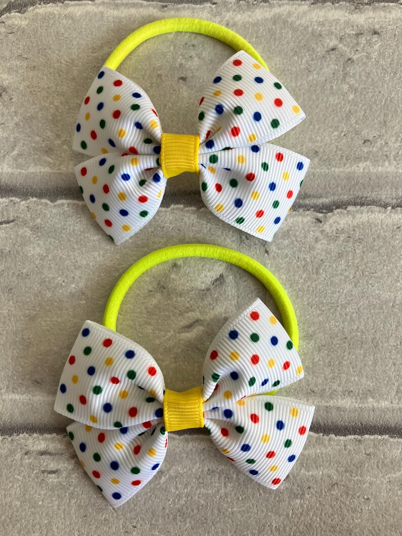 May include: Two neon yellow hair ties with white bows featuring colorful polka dots. The bows have a yellow center.