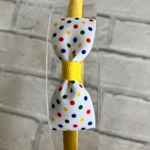 May include: A yellow headband with a white bow decorated with colorful polka dots. The bow has red, yellow, blue, and green polka dots.