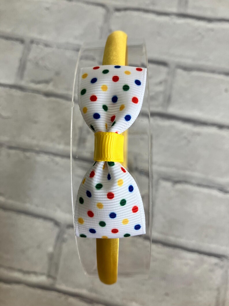 May include: A yellow headband with a white bow decorated with colorful polka dots. The bow has red, yellow, blue, and green polka dots.