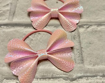 Puede incluir: Dos gomas para el pelo con lazos hechos de un material rosa claro, iridiscente y texturizado. Cada lazo tiene una banda central del mismo tejido brillante con patrón de escamas, unido a una fina goma elástica rosa a juego.