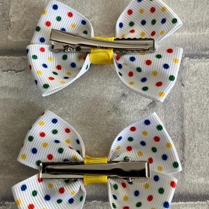 May include: Two white hair bows with a yellow center and colorful polka dots. Each bow has a silver alligator clip attached.