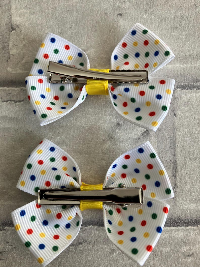 May include: Two white hair bows with a yellow center and colorful polka dots. Each bow has a silver alligator clip attached.