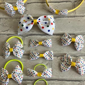 May include: A set of hair accessories featuring white bows with colorful polka dots. The set includes two headbands, four hair ties, and three smaller bows. The bows are all tied with yellow ribbon.