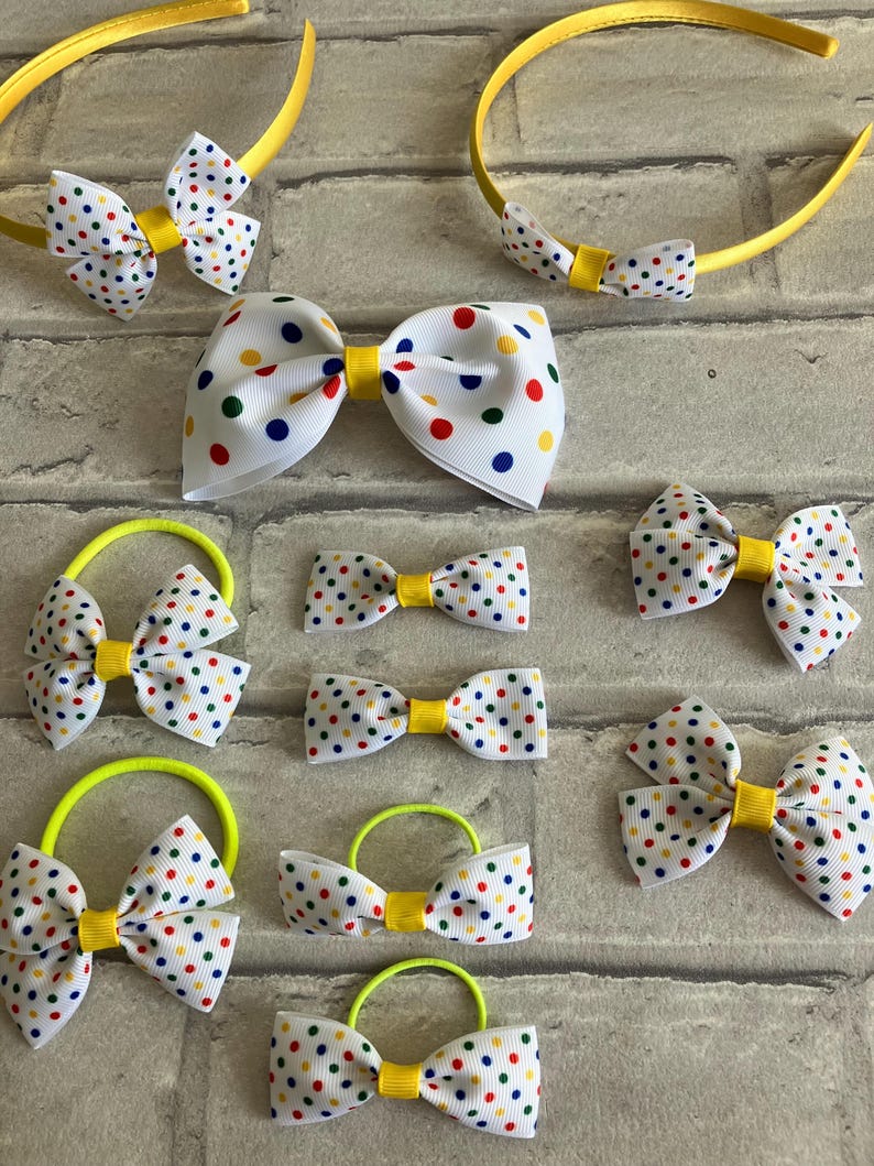 May include: A set of hair accessories featuring white bows with colorful polka dots. The set includes two headbands, four hair ties, and three smaller bows. The bows are all tied with yellow ribbon.