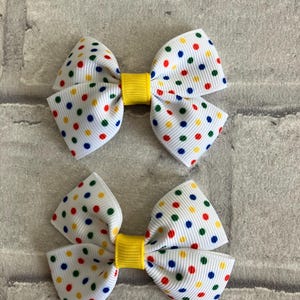 May include: Two white hair bows with a yellow center and multi-colored polka dots. The polka dots are red, blue, green, and yellow.